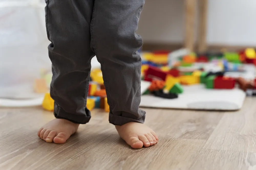 Kind steht barfuß auf einem Holzboden, im Hintergrund liegt Spielzeug